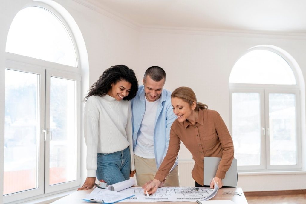 Three people reviewing architectural plans in a bright room with large windows